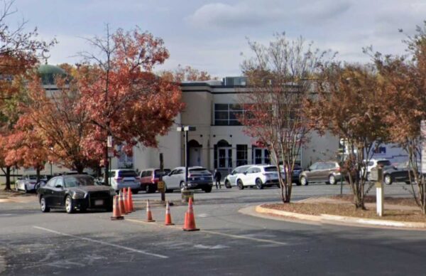 Parking lot with orange construction cones in foreground and a beige building with glass doors behind fall-colored trees.
