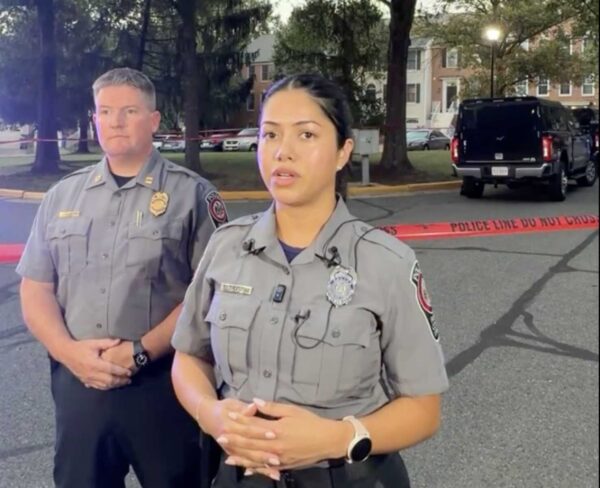 Two police officers with grey uniform shirts stand in front of a red tape line in a parking lot.
