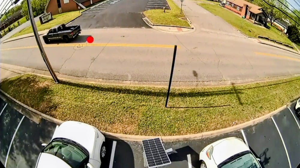 Still from a video capture of electronic monitoring for excessive vehicle noise (via Blue Lines Solutions video) | FFXnow Wide-angle security camera view of a street and parking lot: a black pickup sits near the curb, with a grassy median and a double yellow line across the road in the distance.