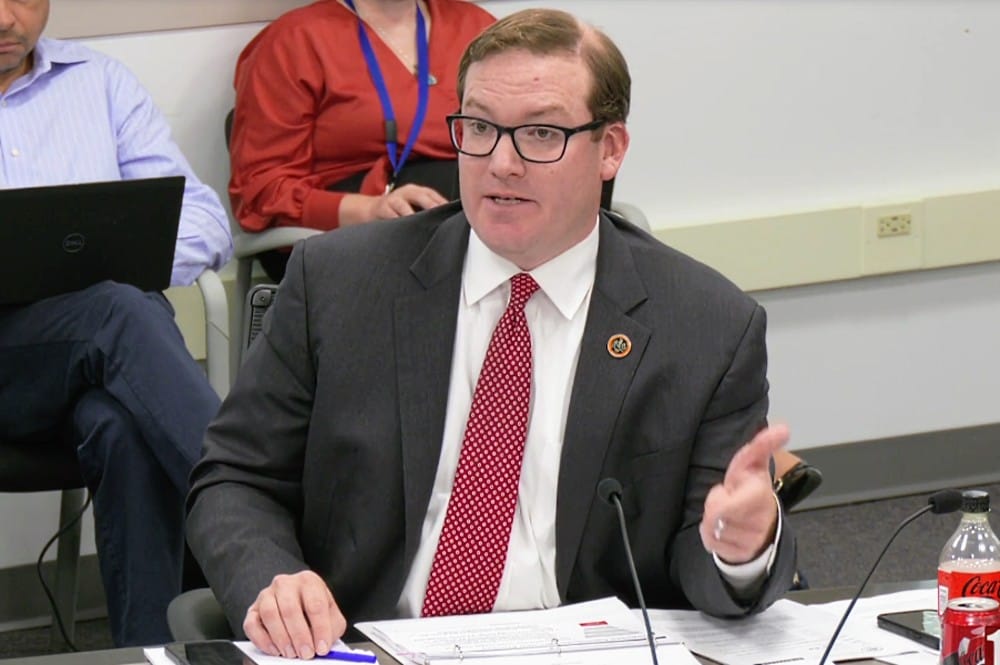 Supervisor Jimmy Bierman discusses changes to Contributory Fund (screenshot via Fairfax County) | FFXnow Man in a gray suit and red tie speaks at a meeting table, gesturing with his right hand.