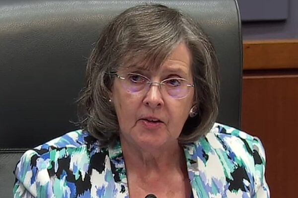 Older woman with short gray hair and glasses speaks at a microphone in a formal meeting room, wearing a blue-green abstract blazer.
