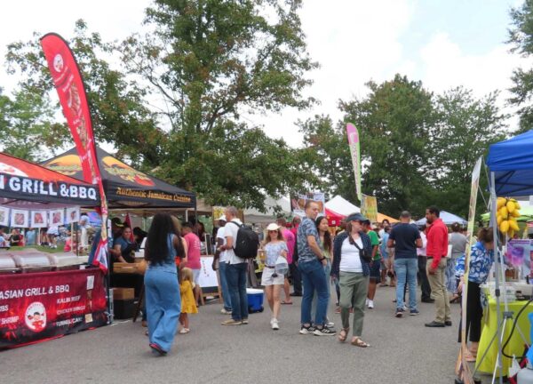 Crowd at an outdoor street fair with colorful vendor tents selling food; banners advertise Asian grill, BBQ, and Jamaican cuisine in the background.