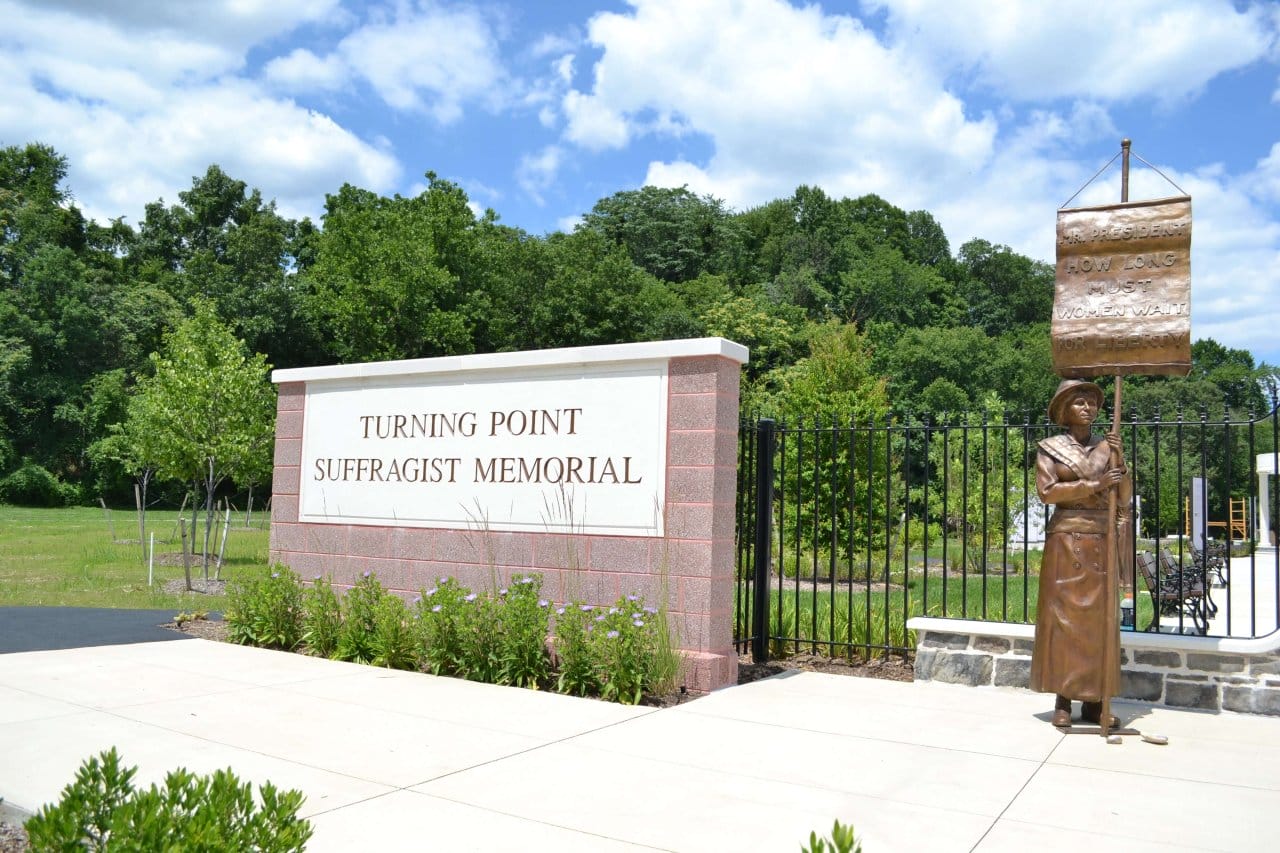 Sculpture of a suffragist holding a banner beside a memorial sign that reads 'Turning Point Suffragist Memorial' at a landscaped park entrance