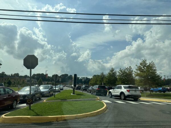 Cars line up in a parking lot around a grassy circular island with a yellow curb, under a blue sky with clouds and power lines above.