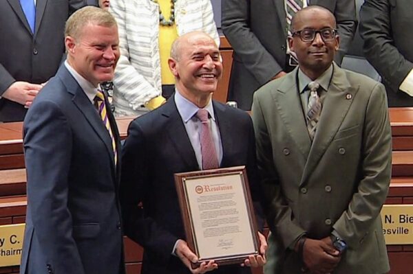 Three men in suits pose for a group photo; the man in the center holds a framed resolution document and smiles, with colleagues on either side in a formal chamber setting.