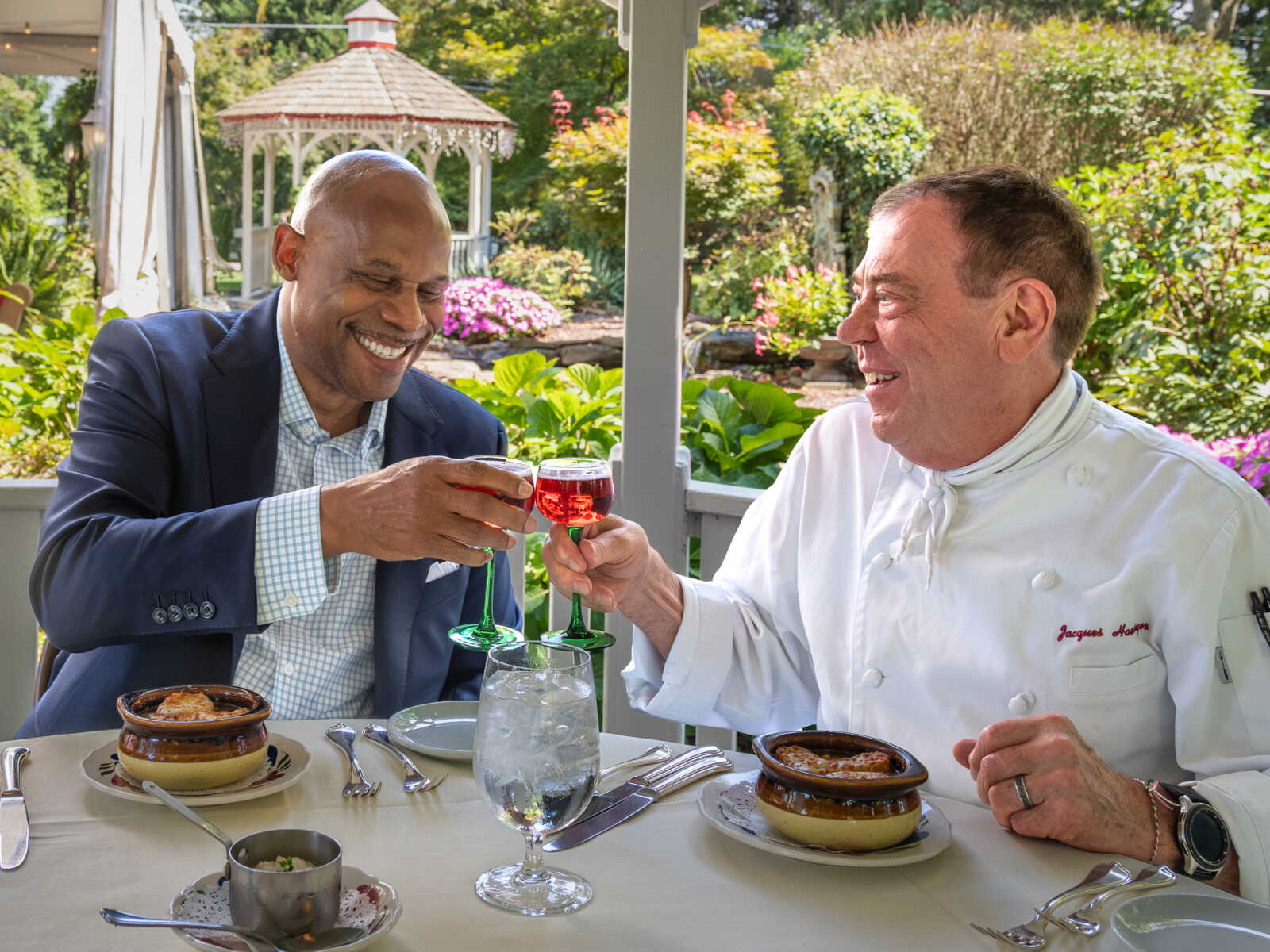 Two men toast with red wine glasses at an outdoor restaurant, one in a navy suit and the other a chef in white, with a garden backdrop.