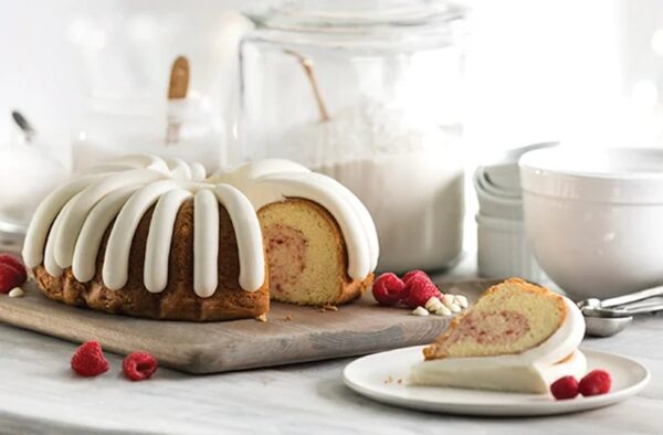 Bundt cake with white icing drizzled over top on a wooden board, sliced to reveal a pink center, with raspberries nearby on a light kitchen counter.