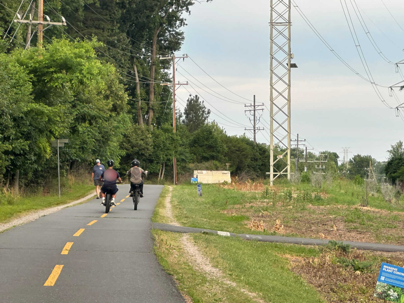 Electric dirt bike riders on the Washington & Old Dominion Trail in Vienna (staff photo by Angela Woolsey) | FFXnow Three cyclists ride along a paved rural bike path with trees on the left and power lines on the right.