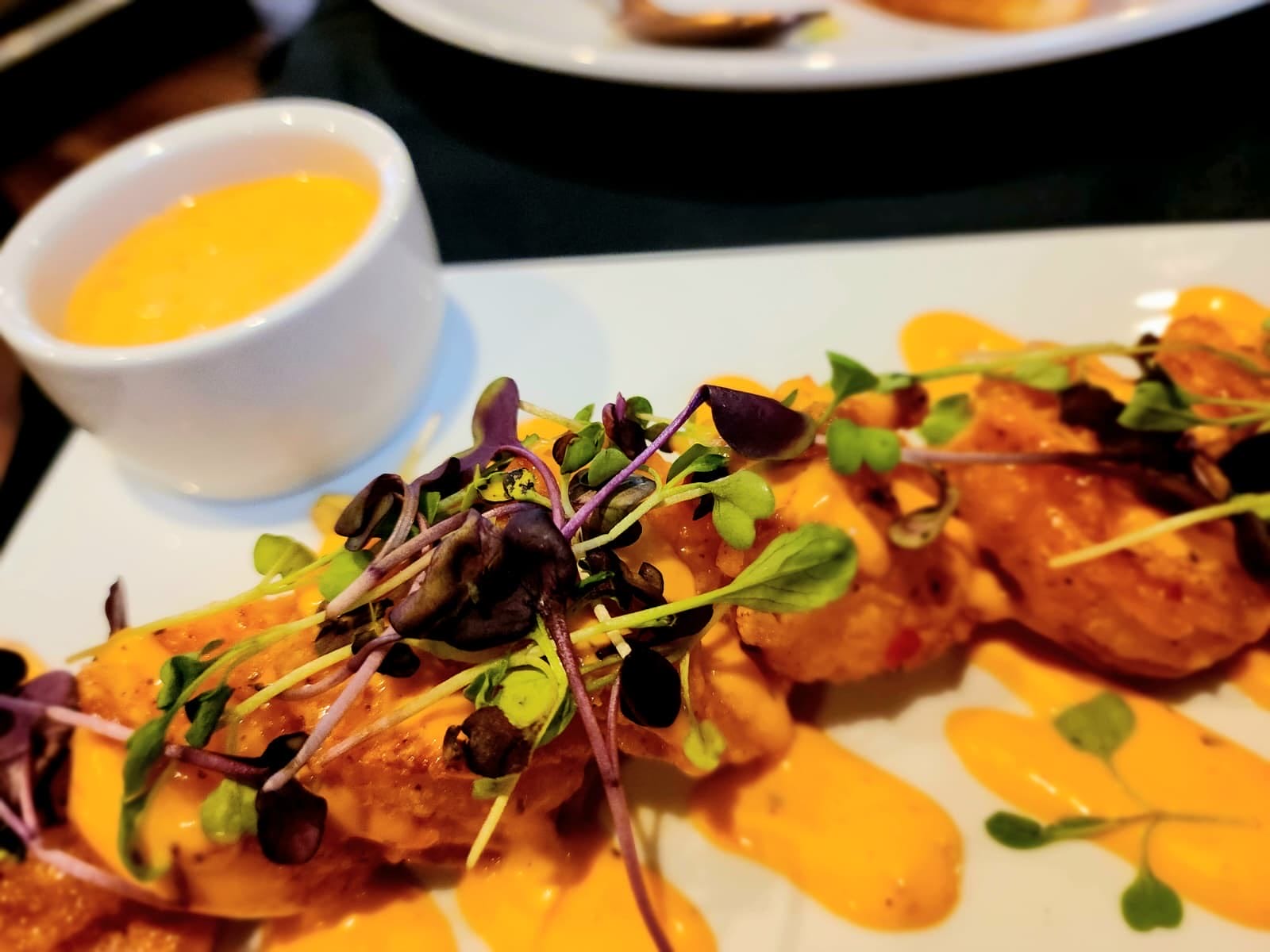 Plate of fried chicken with orange sauce, topped with microgreens and herbs, with a side cup of creamy sauce nearby.
