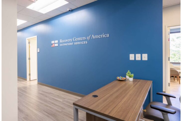 Reception area of an outpatient clinic with a blue wall and a sign reading 'Recovery Centers of America Outpatient Services', wooden desk, small plant, and a chair nearby