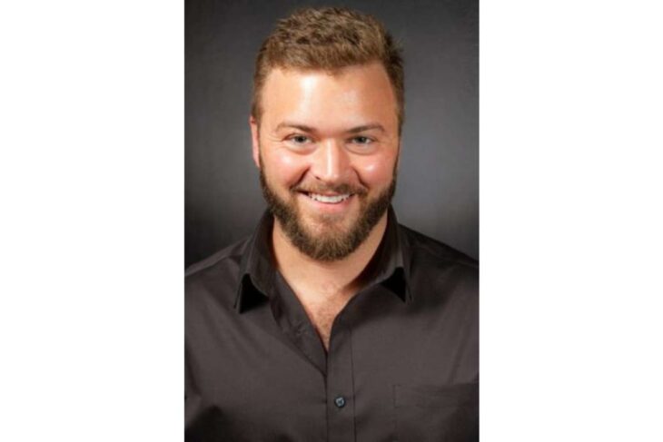 Portrait head-and-shoulders of a smiling man with a beard wearing a black button-down shirt against a dark gray backdrop.