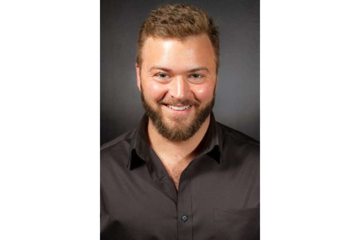 Portrait head-and-shoulders of a smiling man with a beard wearing a black button-down shirt against a dark gray backdrop.