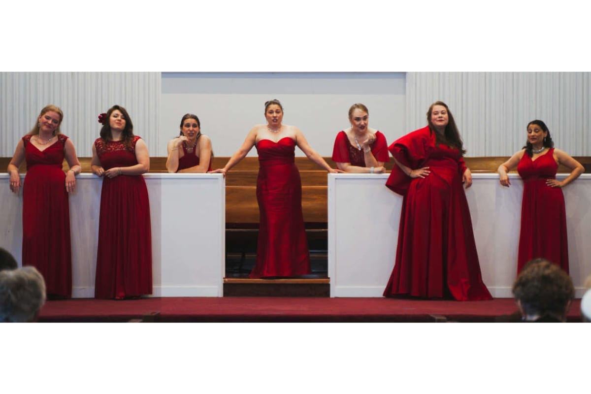 Six women in red formal gowns stand along white podiums on a stage, posing for a photo.