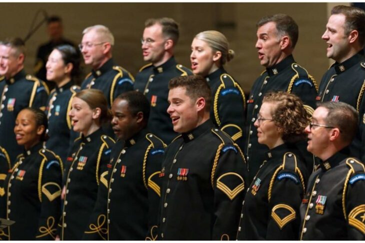 Group of diverse soldiers in black dress uniforms with gold trim singing in a military choir on stage, medals visible on chests.