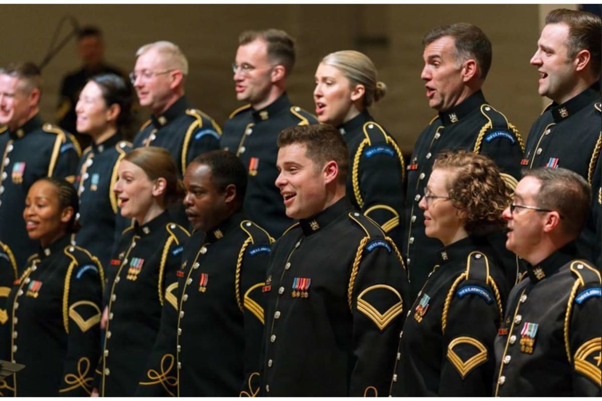 Group of diverse soldiers in black dress uniforms with gold trim singing in a military choir on stage, medals visible on chests.