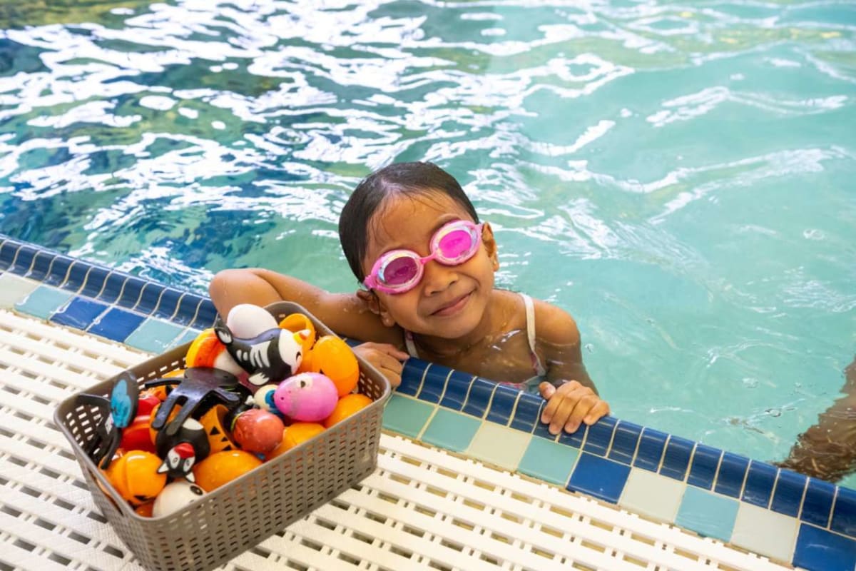 Young girl with pink swim goggles smiles at the pool edge beside a basket of colorful pool toys with stuffed animals and balls in the water nearby.