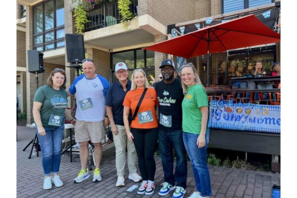 Six adults with race bibs posing for a group photo at an outdoor community event by a brick building and stage setup.