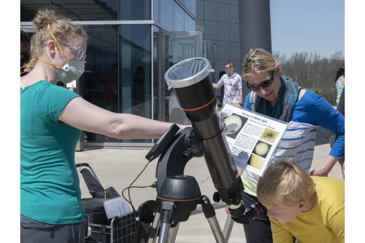 Group of people outside a modern glass-front building examining a large telescope on a tripod.