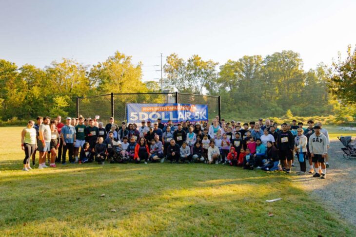 Group photo of runners and families posing on a sunny field behind a banner that reads '5 Dollar 5K' during a community run event.