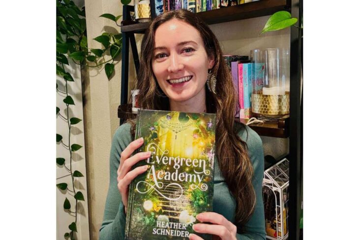 Smiling woman holds a green fantasy novel Evergreen Academy by Heather Schneider in front of a bookshelf.