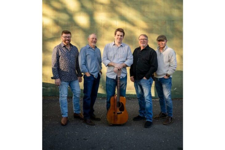 Five men posing outdoors in front of a green wall, with an acoustic guitar standing upright between the center man’s hands.