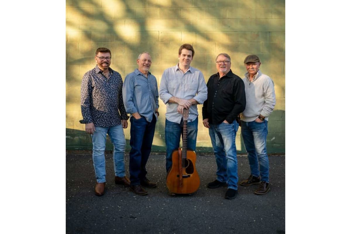 Five men posing outdoors in front of a green wall, with an acoustic guitar standing upright between the center man’s hands.