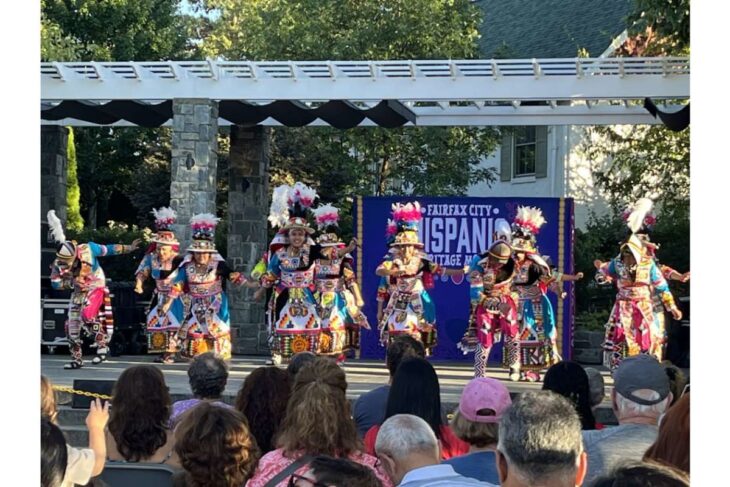 Dancers in bright, ornate costumes with feathered headdresses perform on an outdoor stage while a seated audience watches; banner behind them reads Hispanic Heritage.