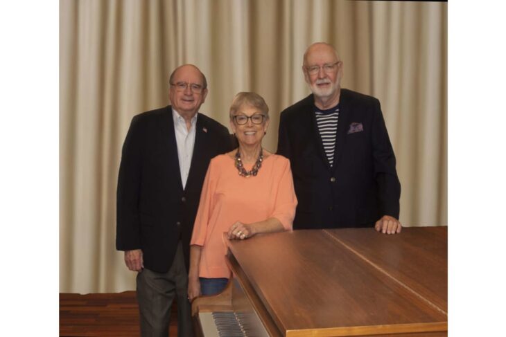 Three smiling seniors posing beside a grand piano in front of beige curtains.
