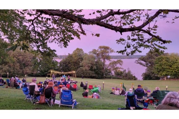 Audience seated on lawn chairs at an outdoor concert on a grassy hill overlooking a lake, with a small stage and string lights at sunset.