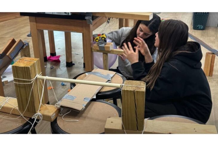 Two students in a workshop collaborate on a hands‑on wood-and-string project, using wooden posts, string, and taped cardboard.