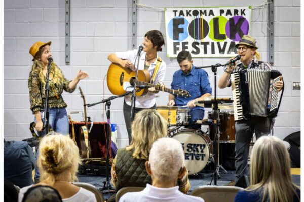 Band on stage at Takoma Park Folk Festival: female vocalist with hat, guitarist, drummer, and accordionist perform as audience watches nearby.
