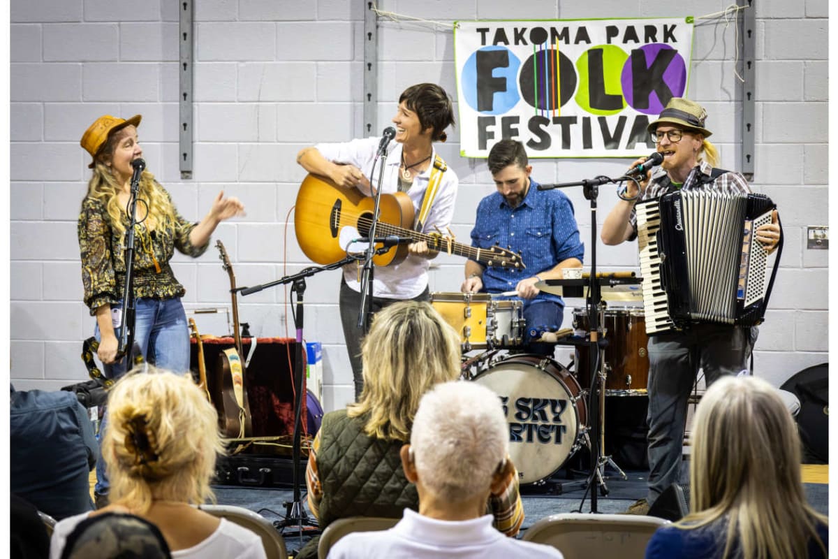Band on stage at Takoma Park Folk Festival: female vocalist with hat, guitarist, drummer, and accordionist perform as audience watches nearby.