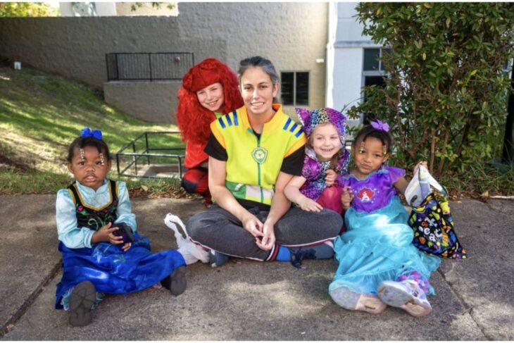 Group of five children in colorful costumes sitting on a sidewalk with a smiling adult in a bright yellow vest outdoors.