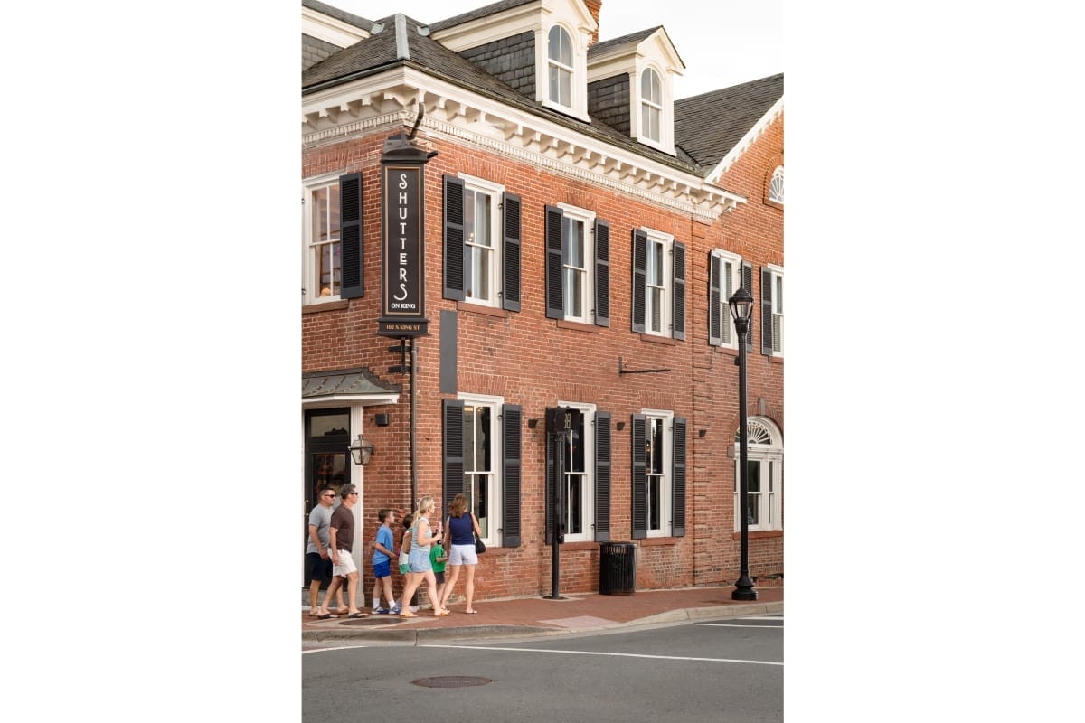 Group of people walking along a brick townhouse with black shutters and a vertical 'Shutter's on King' sign outside.