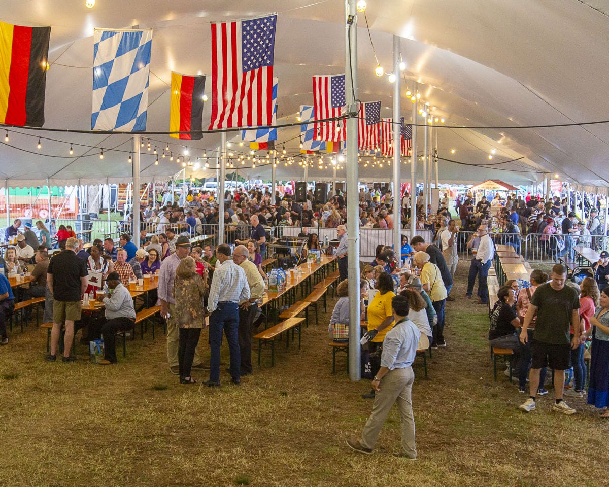 Crowd of people seated at long picnic tables under a large event tent with string lights and flags overhead (American and other flags).