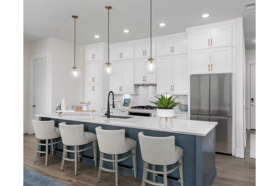Modern kitchen with white cabinets, marble island, blue island base, pendant lights, and stainless steel refrigerator; five beige stools along the island.
