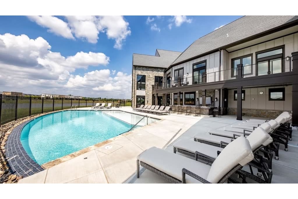 Outdoor hotel pool area with white lounge chairs along a curved pool under a blue sky and clouds, bordered by a fence and modern building.