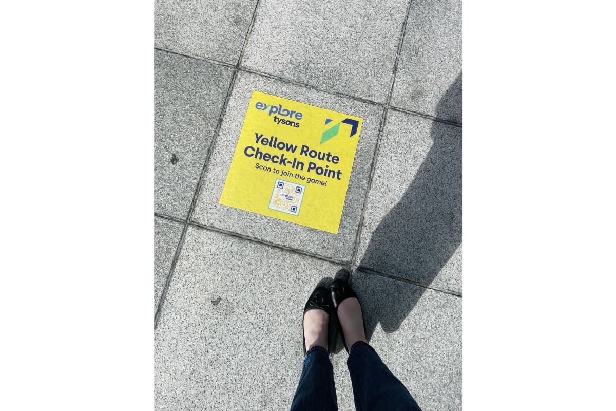 Yellow Route Check-In Point sticker on a concrete sidewalk; person in black flats and jeans stands nearby, with shadow visible. Text on sticker says to scan to join the game.