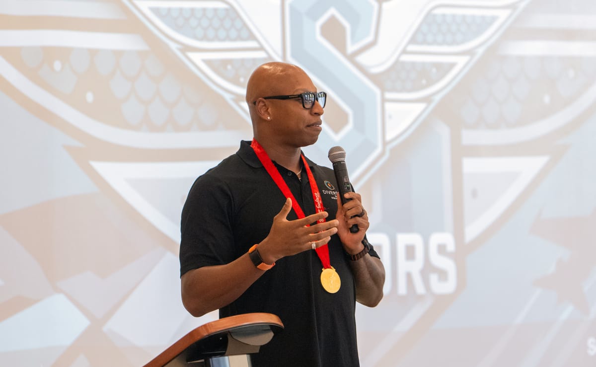 Male presenter wearing a black polo and gold medal on a red ribbon speaks into a handheld microphone at a podium, with a large event backdrop behind him.