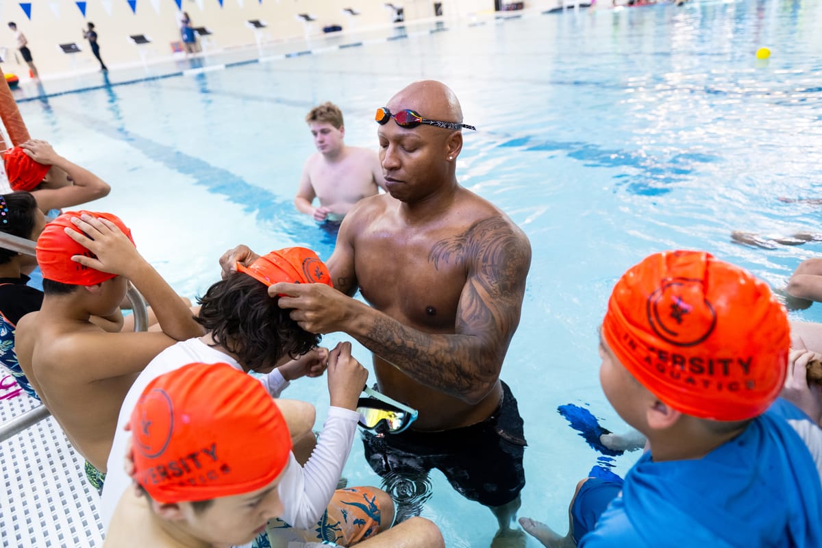 Swim coach adjusting a child’s orange cap as several kids wait at the pool edge for a lesson on a sunny day poolside