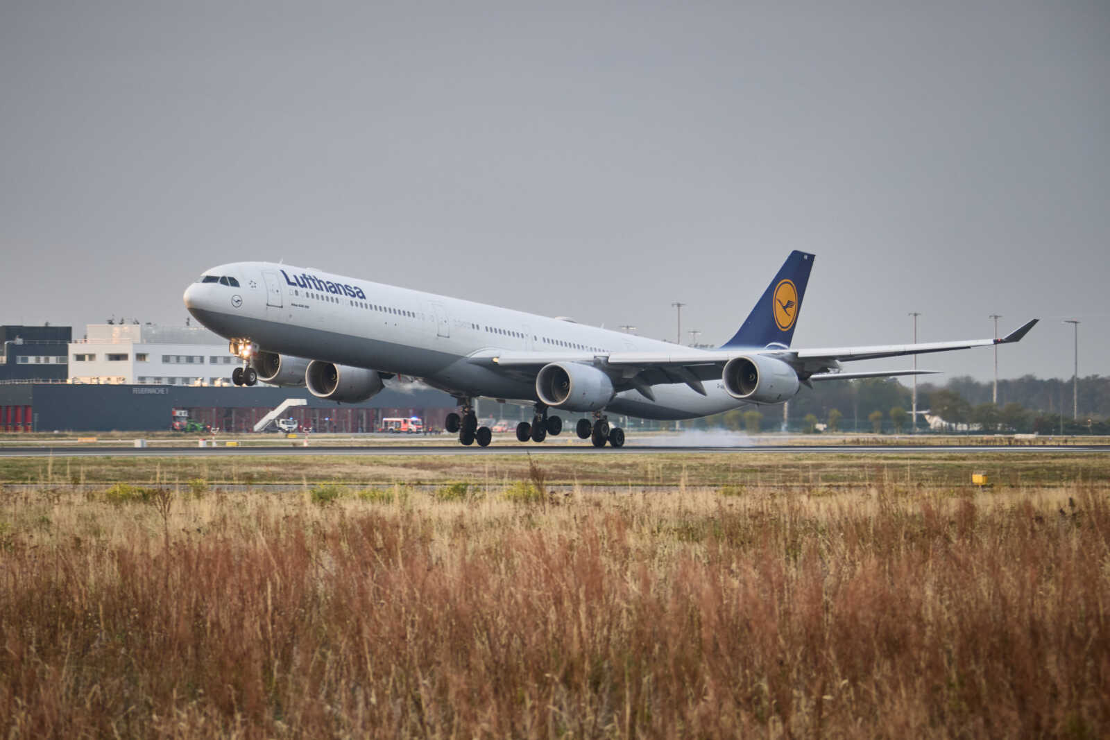 Lufthansa four-engine wide-body jet taking off from a runway with gear down and airport buildings in the background