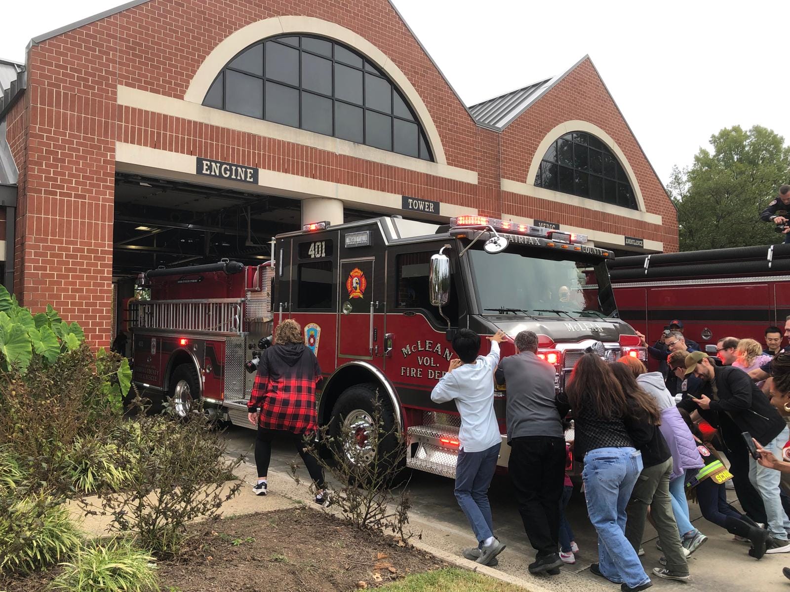 Crowd of people gathered around a large red fire engine parked outside a brick fire station with arched windows.