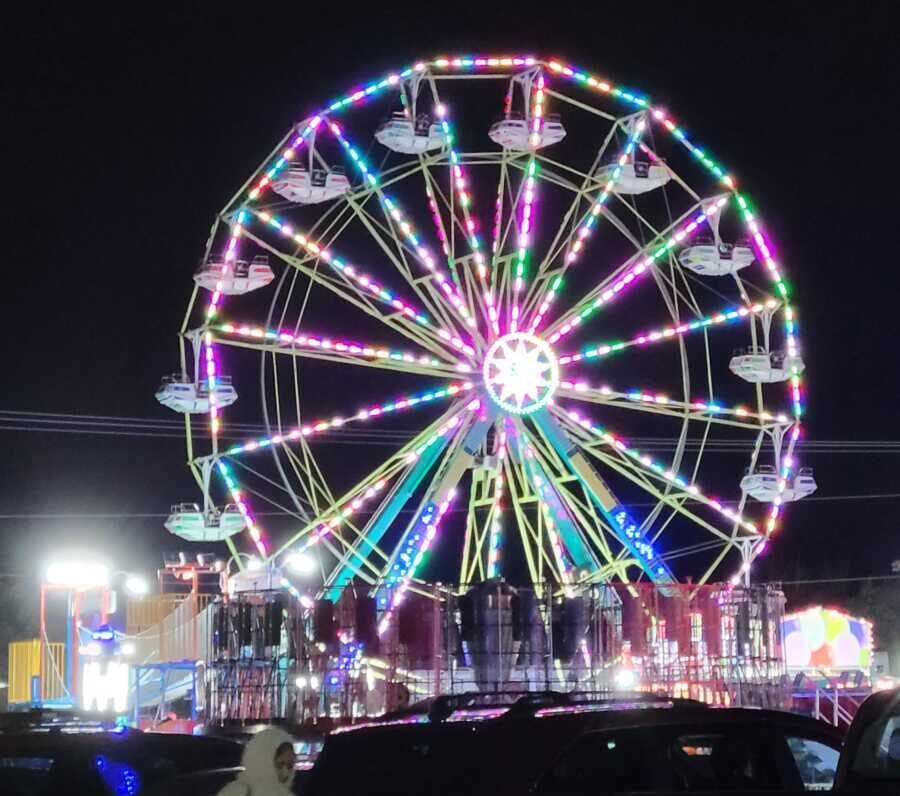 Nighttime carnival with a large multicolored Ferris wheel glowing against the dark sky, with rides and cars in the foreground