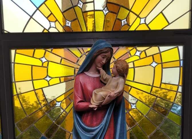 Statue of Mary holding baby Jesus in front of a colorful stained-glass window, placed on a tiled circular planter with green ivy.