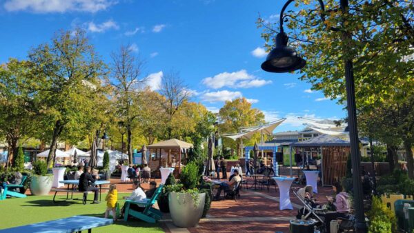 Outdoor plaza with tables, chairs, tents, and umbrellas under a bright blue sky, surrounded by trees and planters while people relax and socialize.