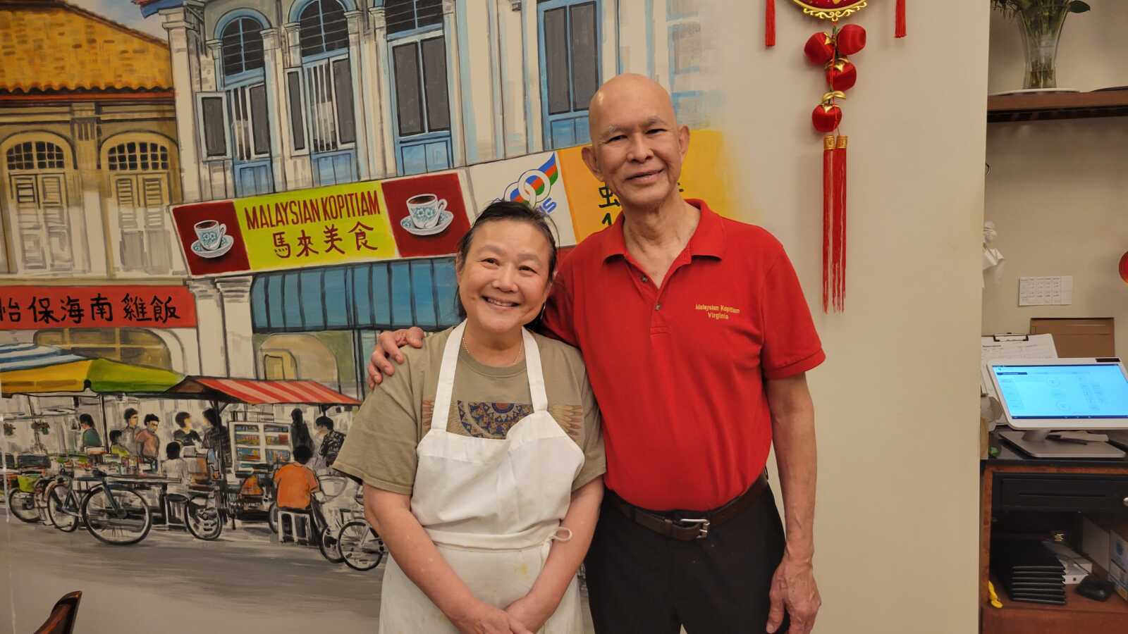 A woman wearing an apron (left) and a man in a red polo shirt stand in front of a colorful mural.
