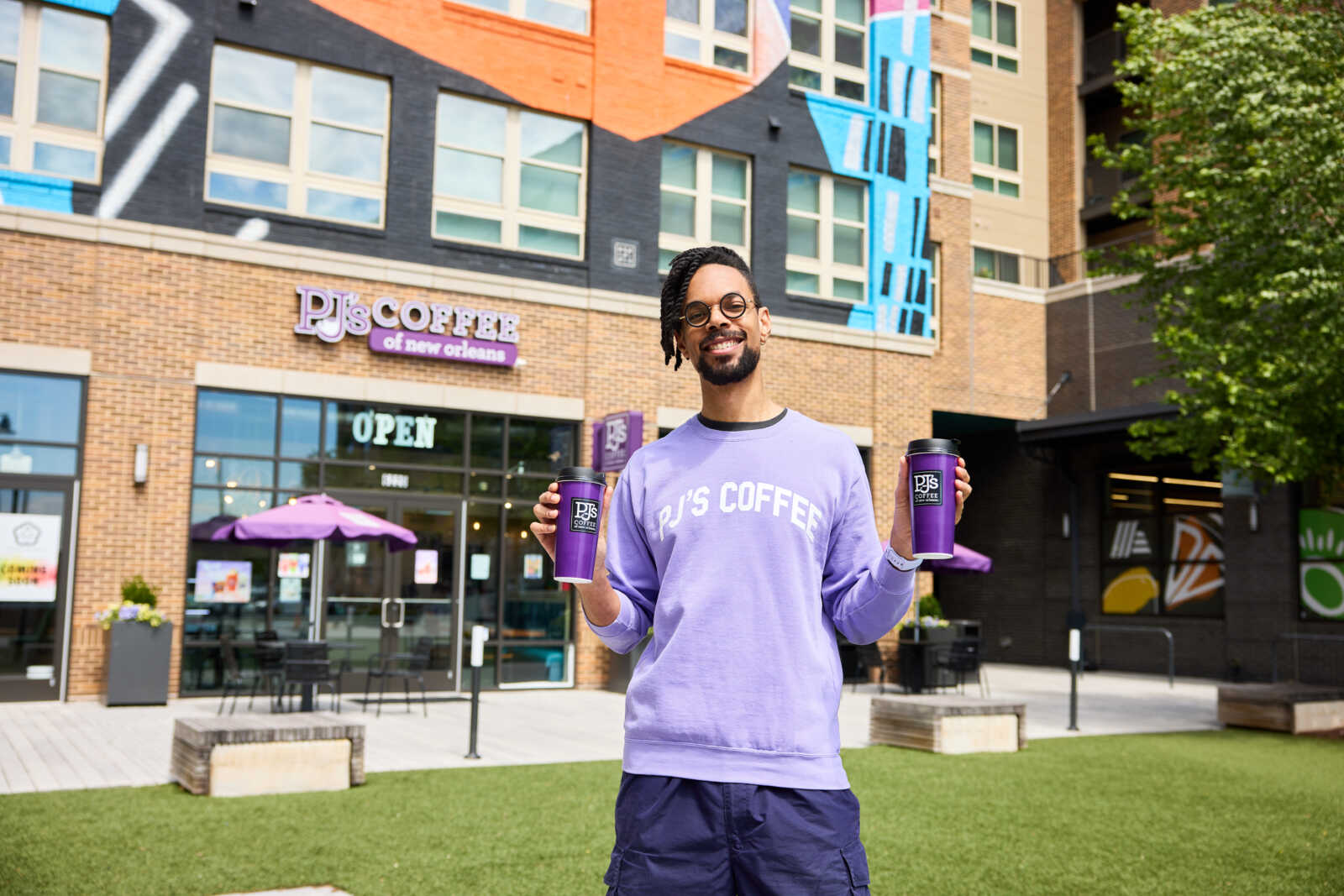 Promotional photo for PJ's Coffee of New Orleans taken as part of the ONE Richmond Highway campaign (courtesy Southeast Fairfax Development Corporation) | FFXnow Smiling man in a lavender PJ's Coffee sweatshirt holds two purple cups outdoors in front of a cafe with colorful mural and outdoor seating.