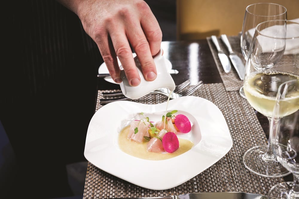 Chef finishes a fine-dining plated dish by pouring light sauce over it, featuring pink radish slices and greens on a white plate.