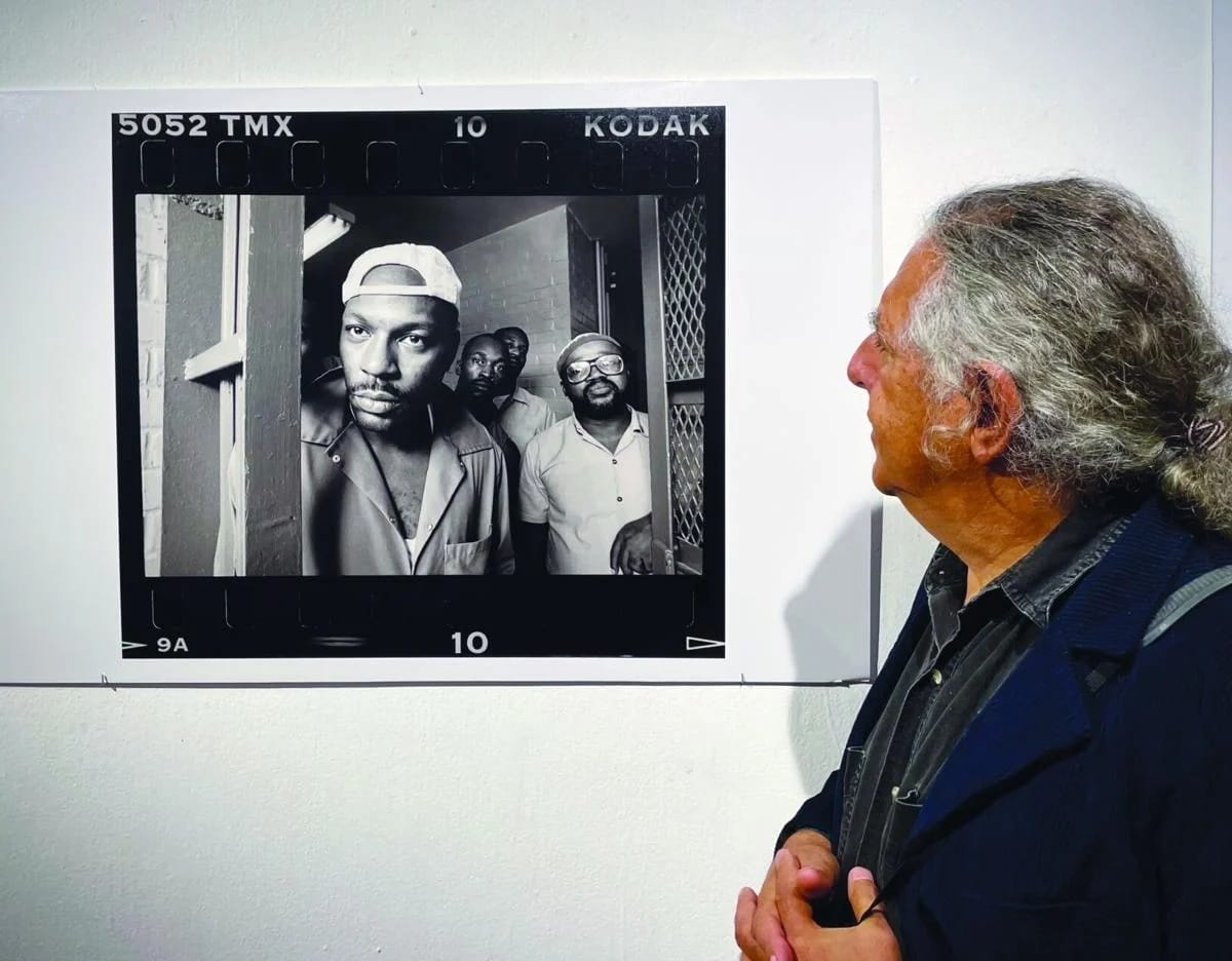 Photographer Lloyd Wolf at the Workhouse Arts Center for an exhibit of his photographs documenting the Lorton Prison's substance abuse rehabilitation program (photo by Aaron Gebresellasie/Inside NoVA) | FFXnow Older man with long gray hair in a dark jacket gazes at a wall-mounted black-and-white photo depicting four men in a doorway.