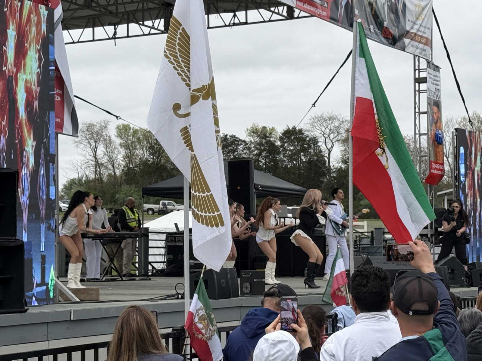 Dance performance on an outdoor stage with female dancers in white outfits and boots, Italian flag visible in the foreground and a large stage screen on the side.